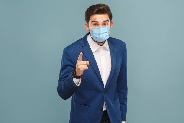 Finaly warning. Serious man with surgical medical mask pointing finger and alert you. Business people medicine and health care concept. Indoor, studio shot on blue background - Фото, изображение