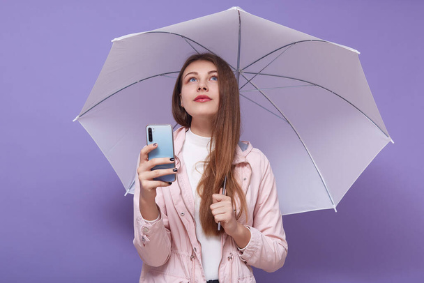 Portrait of pensive good looking tender female looking up, holding smartphone and pink umbrella, using her device, taking photos, having long hair, staying alone, being dreamy. Youth concept. - Φωτογραφία, εικόνα