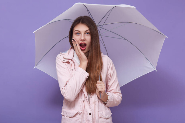 Indoor studio shot of emotional shocked young lady opening mouth and eyes widely, putting one hand on cheeck, holding umbrella, finding out news, being surprised. People and emotions concept. - Fotoğraf, Görsel