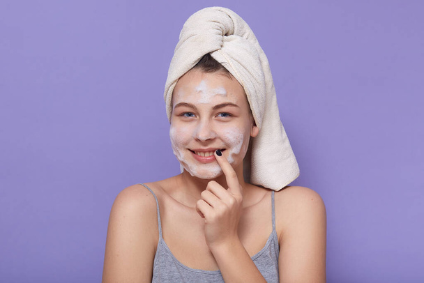 Picture of cheerful tender young girl having spa procedures, wearing pajama, having towel on her wet hair, looking directly at camera, smiling sincerely, applying mask on face. Care concept. - Foto, Imagem