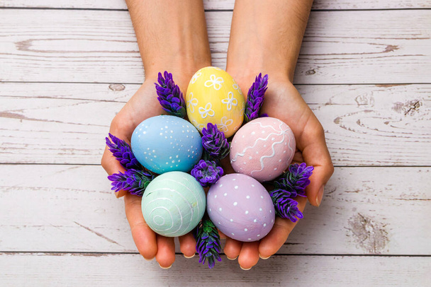 easter concept. closeup beautiful woman hands holding hand-painted easter eggs in tender pastel colors and lavender flowers over wooden table - Photo, Image