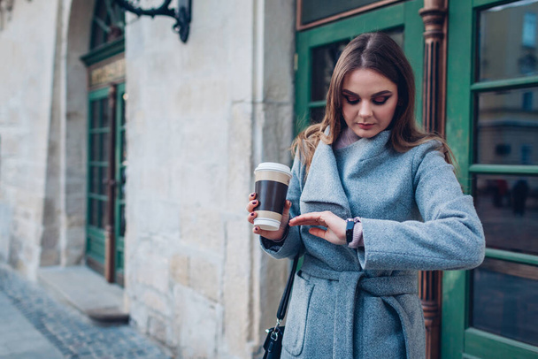 Молодая стильная женщина, глядя на ручные часы, пьет кофе на городской улице в ожидании друзей. Весенняя мода
 - Фото, изображение