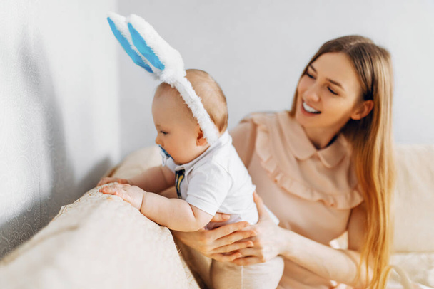 Mom and baby with rabbit ears, with Easter eggs in their hands, sitting on the sofa, parents and children play indoors. Family celebrates Easter - Zdjęcie, obraz