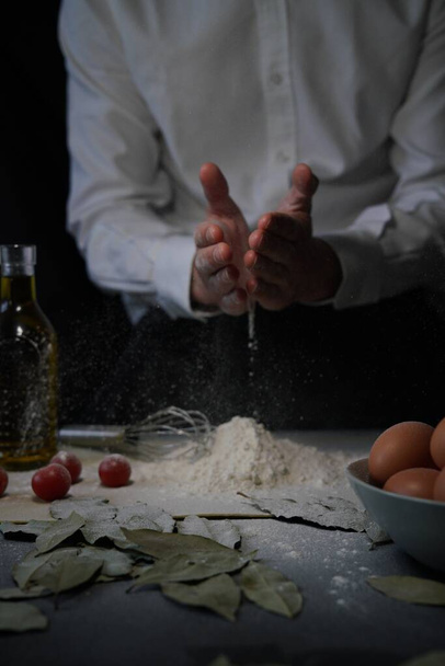 A man pouring flour onto the table next to a bowl of eggs, tomatoes and a bottle - Photo, image