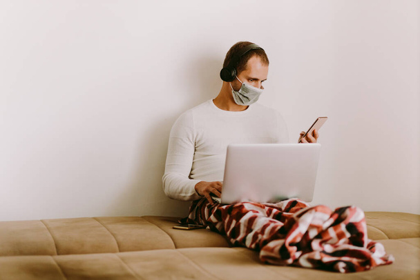 człowiek korzystający z laptopa w domu podczas kwarantanny w domu. Mężczyzna rasy kaukaskiej pracujący zdalnie w domu i noszący maskę przeciwwirusową. - Zdjęcie, obraz