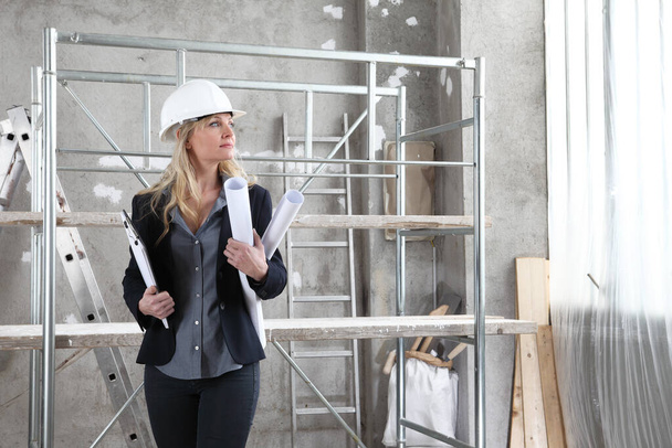 woman architect or construction engineer wear helmet and holds folder and blueprint inside a building site with windows and scaffolding in the background - Photo, Image