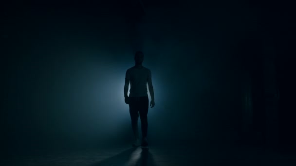 Young man in sports wear walking through smoky studio or scene directly to camera. Backlight, bright spotlight on background. - Footage, Video