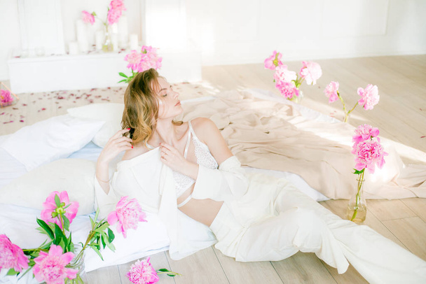 Cute tender young girl with blond hair in white clothes and lace underwear surrounded by pink peonies on the bed on the bed in the studio. Spring and flowers. Beauty and Fashion - Valokuva, kuva