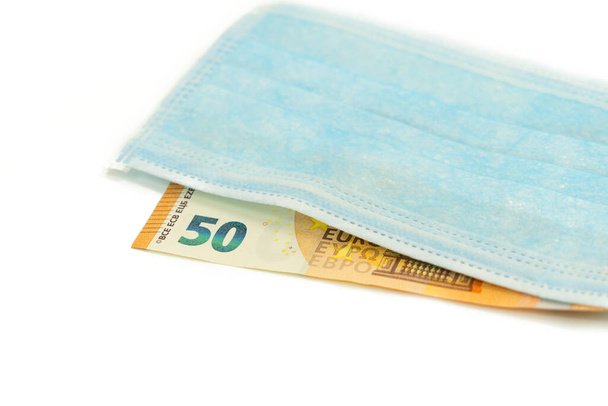 Blue medical mask lies on a banknote of 50 euros. Concept of buying a mask in Europe, protection against the virus. Isolated white background. View from above. - Zdjęcie, obraz