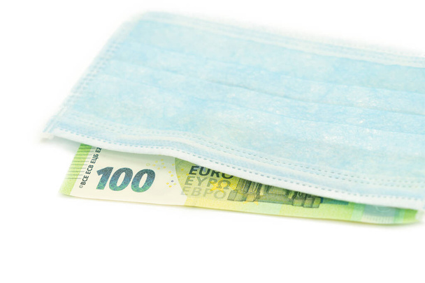 Niebieska maska medyczna leży na banknotze 100 euro. Koncepcja zakupu maski w Europie, ochrona przed wirusem. Odizolowane białe tło. Widok z góry. - Zdjęcie, obraz