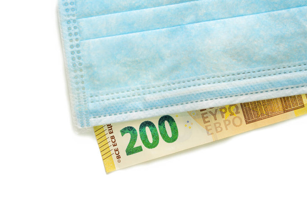 A blue medical mask lies on a banknote of 200 euros. Concept of buying a mask in Europe, protection against the virus. Isolated white background. - Photo, Image