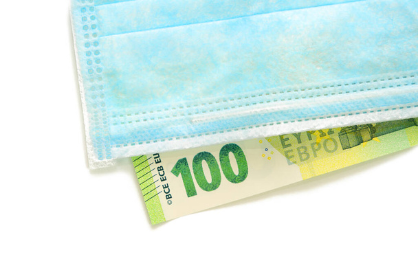 Blue medical mask lies on a banknote of 100 euros. Concept of buying a mask in Europe, protection against the virus. Isolated white background. View from above. - Photo, Image