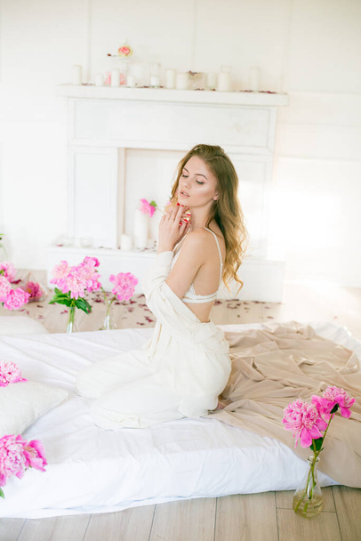 Cute tender young girl with blond hair in white clothes and lace underwear surrounded by pink peonies on the bed on the bed in the studio. Spring and flowers. Beauty and Fashion - 写真・画像
