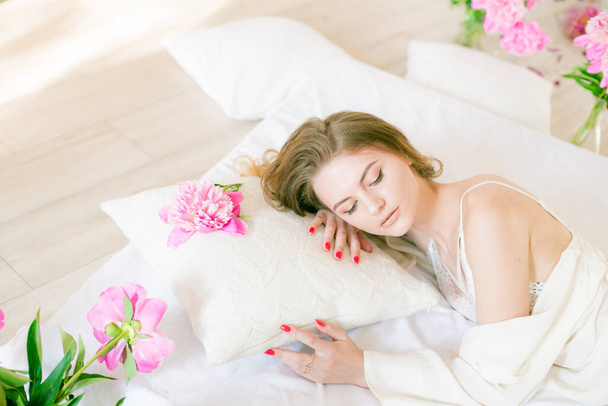 Cute tender young girl with blond hair in white clothes and lace underwear surrounded by pink peonies on the bed on the bed in the studio. Spring and flowers. Beauty and Fashion - Фото, изображение