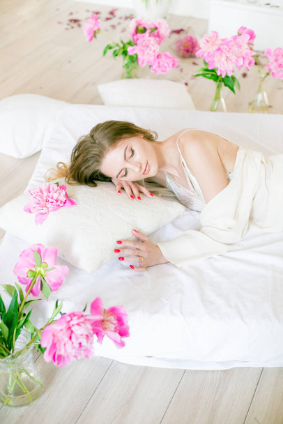 Cute tender young girl with blond hair in white clothes and lace underwear surrounded by pink peonies on the bed on the bed in the studio. Spring and flowers. Beauty and Fashion - 写真・画像