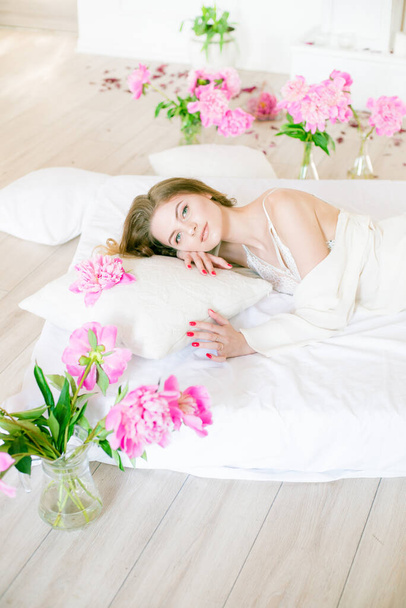 Cute tender young girl with blond hair in white clothes and lace underwear surrounded by pink peonies on the bed on the bed in the studio. Spring and flowers. Beauty and Fashion - Foto, imagen
