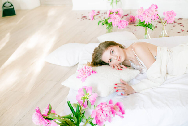 Cute tender young girl with blond hair in white clothes and lace underwear surrounded by pink peonies on the bed on the bed in the studio. Spring and flowers. Beauty and Fashion - Фото, изображение