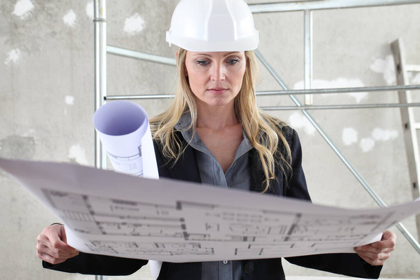 woman architect or construction engineer reading blueprint wear helmet inside a building site with scaffolding in the background - Foto, Imagen