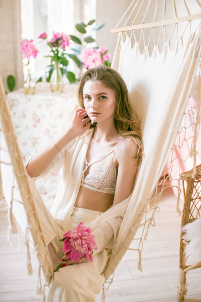 Cute tender young girl with blond long hair surrounded by pink peonies is resting in a hammock in the studio. Spring and flowers. Beauty and Fashion - Foto, Bild