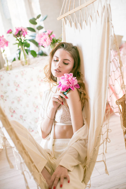 Cute tender young girl with blond long hair surrounded by pink peonies is resting in a hammock in the studio. Spring and flowers. Beauty and Fashion - Photo, Image