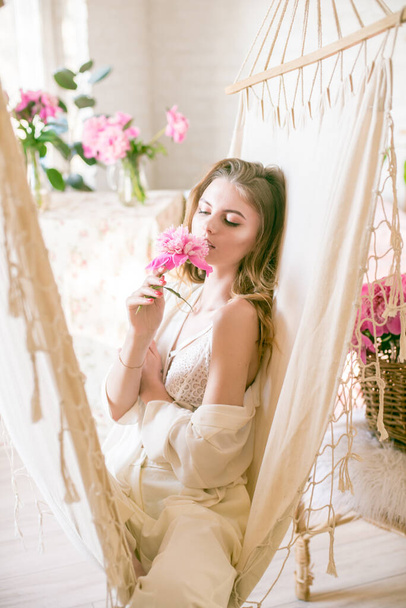 Cute tender young girl with blond long hair surrounded by pink peonies is resting in a hammock in the studio. Spring and flowers. Beauty and Fashion - Fotografie, Obrázek