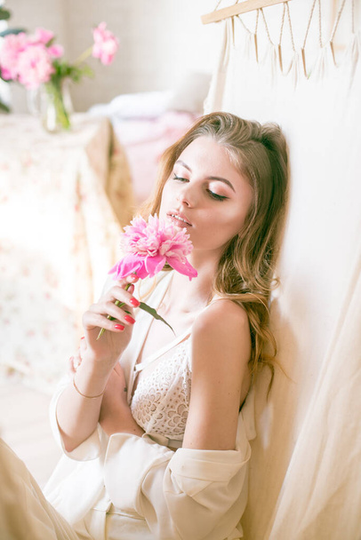 Cute tender young girl with blond long hair surrounded by pink peonies is resting in a hammock in the studio. Spring and flowers. Beauty and Fashion - Photo, image