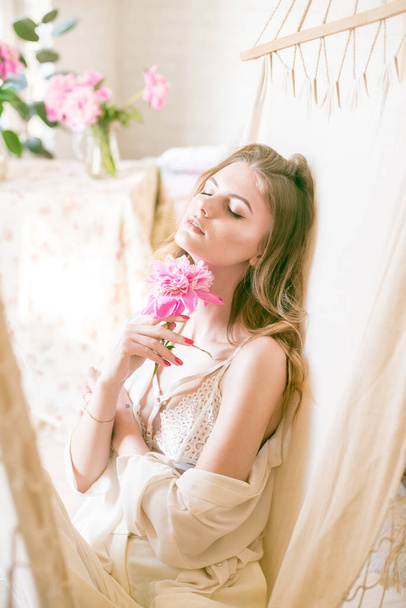 Cute tender young girl with blond long hair surrounded by pink peonies is resting in a hammock in the studio. Spring and flowers. Beauty and Fashion - Foto, afbeelding