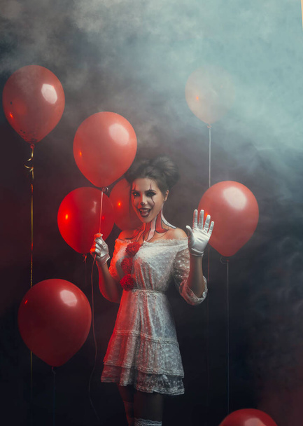 portrait cute young woman crazy smiling face clown in white costume stands backdrop dark gothic room holding red balloons waved hand. art creative bright halloween make-up hairstyle. Fog smoke party  - Photo, Image