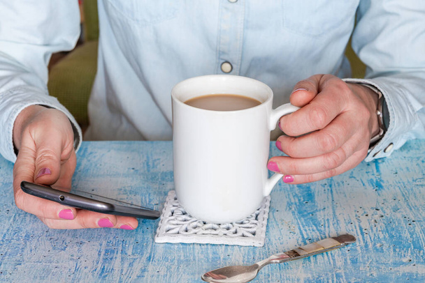 Жіночі руки тримають мобільний телефон і чашку гарячого напою на столі, крупним планом з вибірковим фокусом
 - Фото, зображення