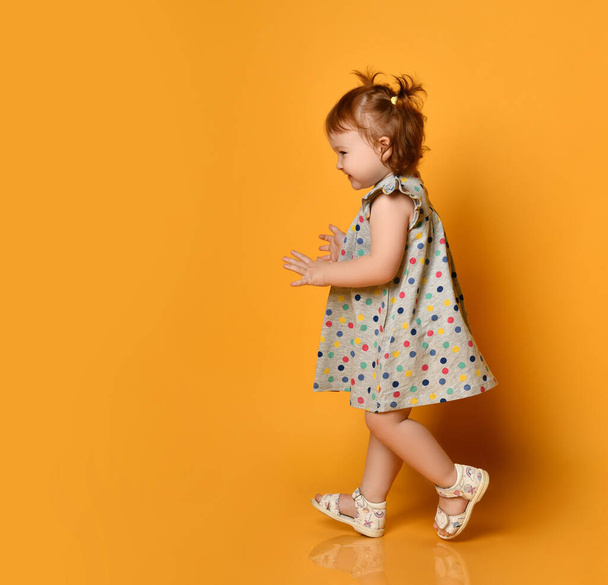 Ginger toddler female with two ponytails, in gray polka dot dress and white sandals. Smiling while posing on orange background - Foto, imagen