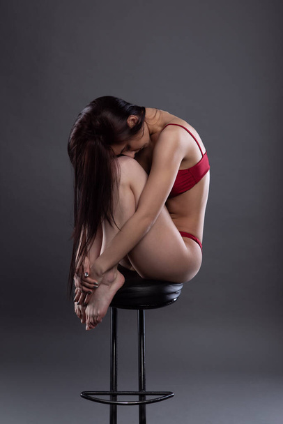 A sexy girl in red underwear sits sadly on a bar stool, a fashion portrait on a gray isolated background - Fotografie, Obrázek