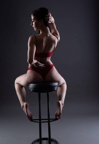 a beautiful young woman in sexy red underwear poses sitting on a chair with her back to the camera - Foto, afbeelding