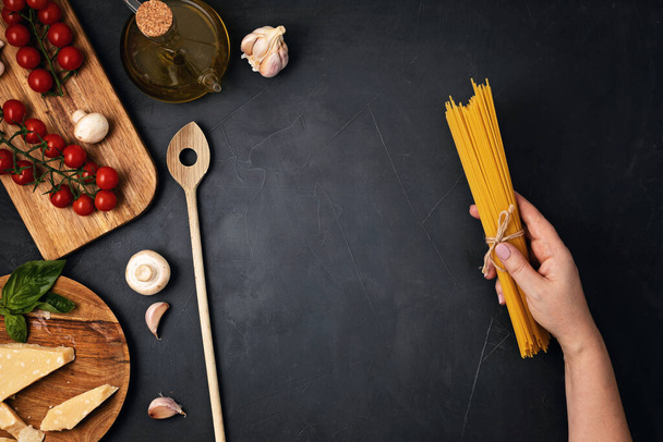 Flat lay of woman hand holding spaghetti and ingredients for cooking italian pasta. Spaghetti, tomatoes, oil, garlic, parmezan. Top view of traditional italian cusine concept - Photo, Image