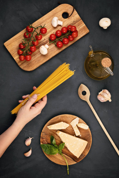 Puesta plana de mano de mujer sosteniendo espaguetis e ingredientes para cocinar pasta italiana. Espaguetis, tomates, aceite, ajo, parmezano. Vista superior del concepto tradicional italiano cusine
 - Foto, Imagen