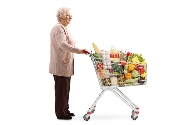 Full length profile shot of an elderly lady with a shopping cart full of food isolated on white background - Valokuva, kuva