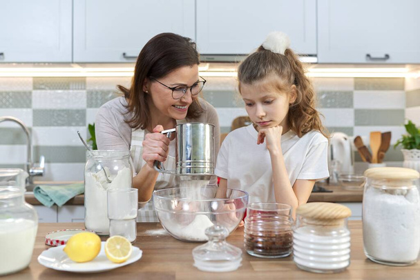 Mother and child preparing bakery together in home kitchen. Woman sifts flour, teaches her little daughter how to cook cupcakes. Homemade food, family, parent child communication - Zdjęcie, obraz