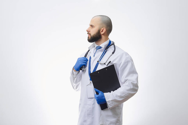 Γιατρός με γάντια μιας χρήσης με στηθοσκόπιο και ντοσιέ. Ένας φαλακρός γιατρός με γενειάδα ετοιμάζεται να εξετάσει ασθενείς.. - Φωτογραφία, εικόνα