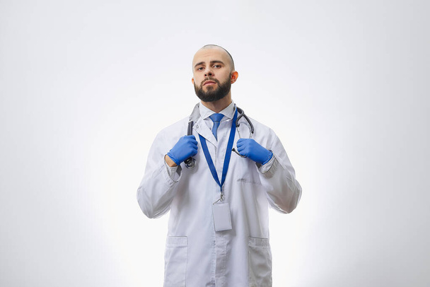 Ένας σοβαρός γιατρός με αναλώσιμα ιατρικά γάντια που κρατάει στηθοσκόπιο. Ένας φαλακρός γιατρός με γενειάδα ετοιμάζεται να εξετάσει έναν ασθενή.. - Φωτογραφία, εικόνα