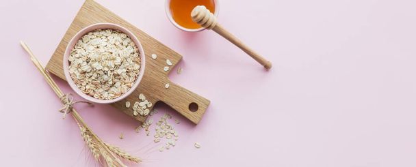Bowl of dry oat flakes with honey and ears of wheat on light pink background. Cooking oats porridge concept. Top view - Photo, image