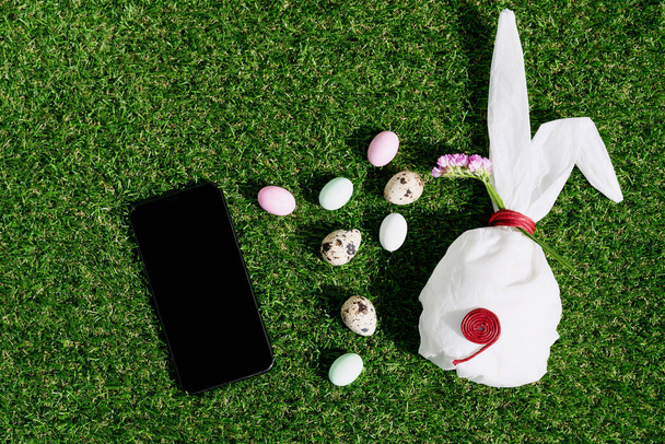 Chocolate Easter eggs glazed with pastel coloring and quail eggs over green grass background. Bunny shaped white baking paper. Black cell phone. Happy Easter concept. Copy space. Top view - Photo, Image