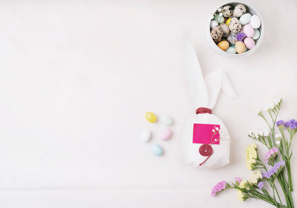 Chocolate Easter eggs glazed with pastel coloring and quail eggs in a bowl and over white background. Bunny shaped white baking paper. The place for the text on the paper. Decorated with spring flowers. Happy Easter concept. Copy space. Top view - Photo, Image