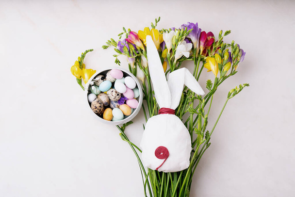 Chocolate Easter eggs glazed with pastel coloring and quail eggs in a bowl over spring flowers on the white background. Bunny shaped white baking paper. Happy Easter concept. Copy space. Top view - Photo, Image