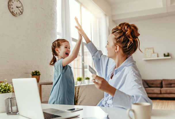 Low angle of excited small daughter giving high five to mother and screaming while celebrating successful online shopping using laptop at table in light living roo - Foto, imagen