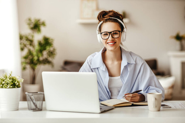 Dedicated female freelancer in glasses and wireless headphones smiling at camera while sitting at laptop with mug of hot drink and writing in notepad in light modern living roo - Photo, Image