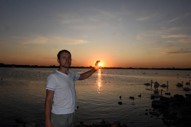 Nuori mies seisoo valtavan järven rannalla valkoisten joutsenten kanssa, hän yrittää saada auringon käteensä, kauniin auringonlaskun.
. - Valokuva, kuva