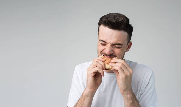 Young man isolated over background. Picture of guy eating burger and biting it. Tasty delisious fatning meal. Eating with pleasure. No guilty for eating bad food. Alone in studio. - Fotó, kép