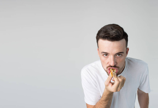 Young man isolated over background. Strange weirdo look straight on camera and biting piece of yummy tasty burger. Guy in white shirt posing alone in studio. Unhealthy lifestyle. - Foto, imagen