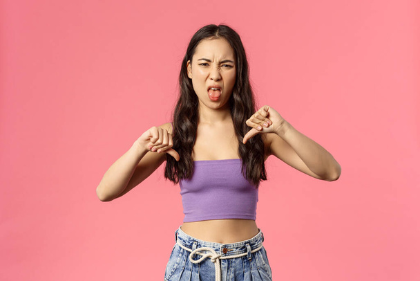 It was really bad. Portrait of disappointed young attractive woman tell friend her date wend awful, show thumbs-down and tongue, grimacing from aversion and dislike, disappointed, pink background - Photo, Image