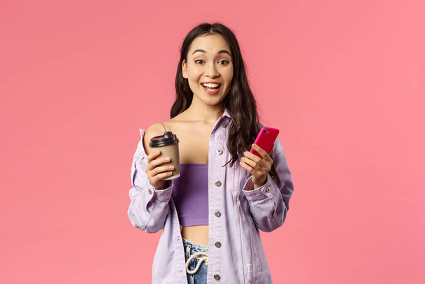 Портрет удивленной привлекательной, улыбающейся женщины, держащей смартфон и кофе на вынос, видящей друга на улице и взволнованной встречей с ней, стоящей на розовом фоне с энтузиазмом
 - Фото, изображение