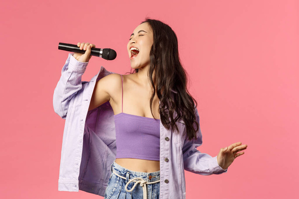 Porträt eines aufgeregten, gut aussehenden, unbeschwerten asiatischen Mädchens, das mit ganzem Herzen singt, sich anstrengen muss, um den höchsten Ton zu erreichen, Karaoke singt, die Augen schließt und es genießt, im Stehen vor rosa Hintergrund aufzutreten - Foto, Bild